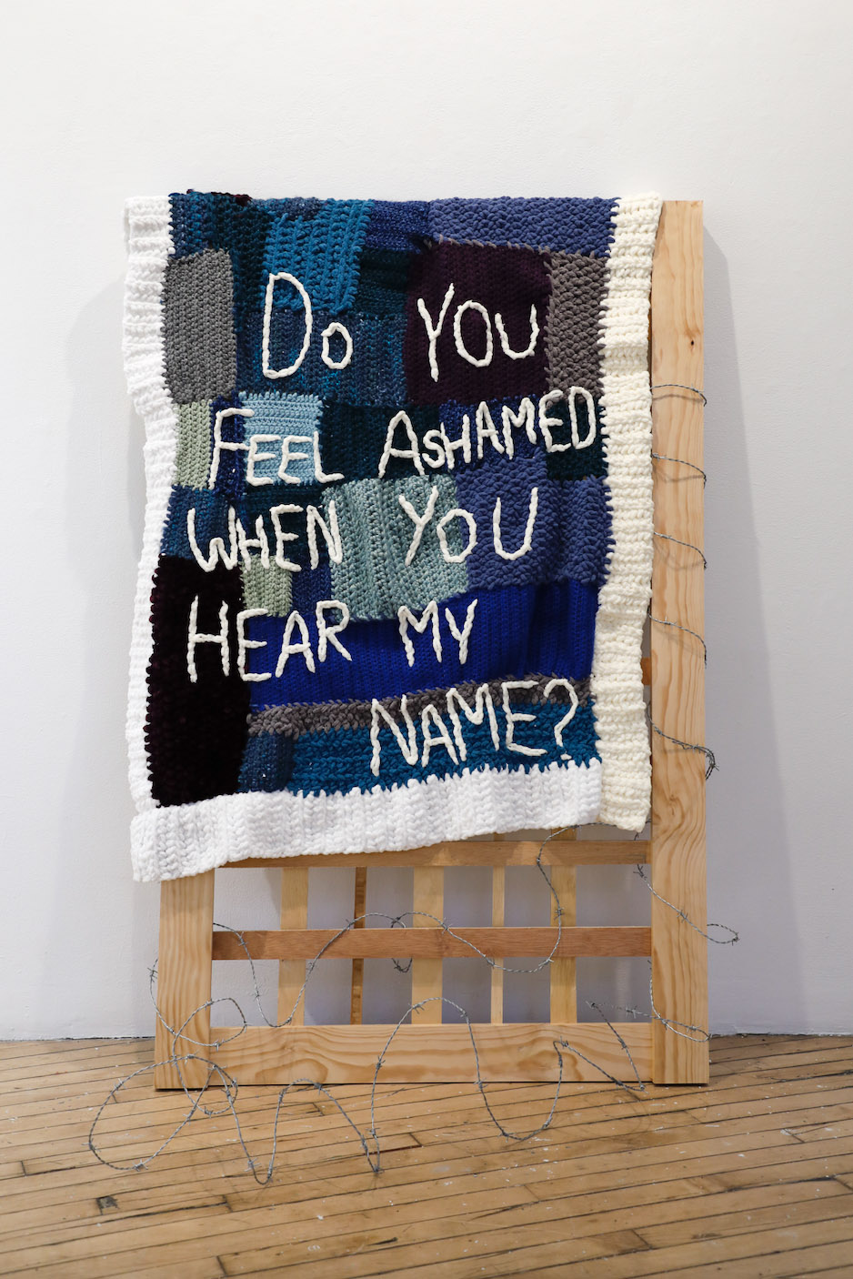Image of crocheted blanket in shades of blue, purple, black, and white yarn over light brown rectangular wooden frame and gray barbed wire at the frame's bottom