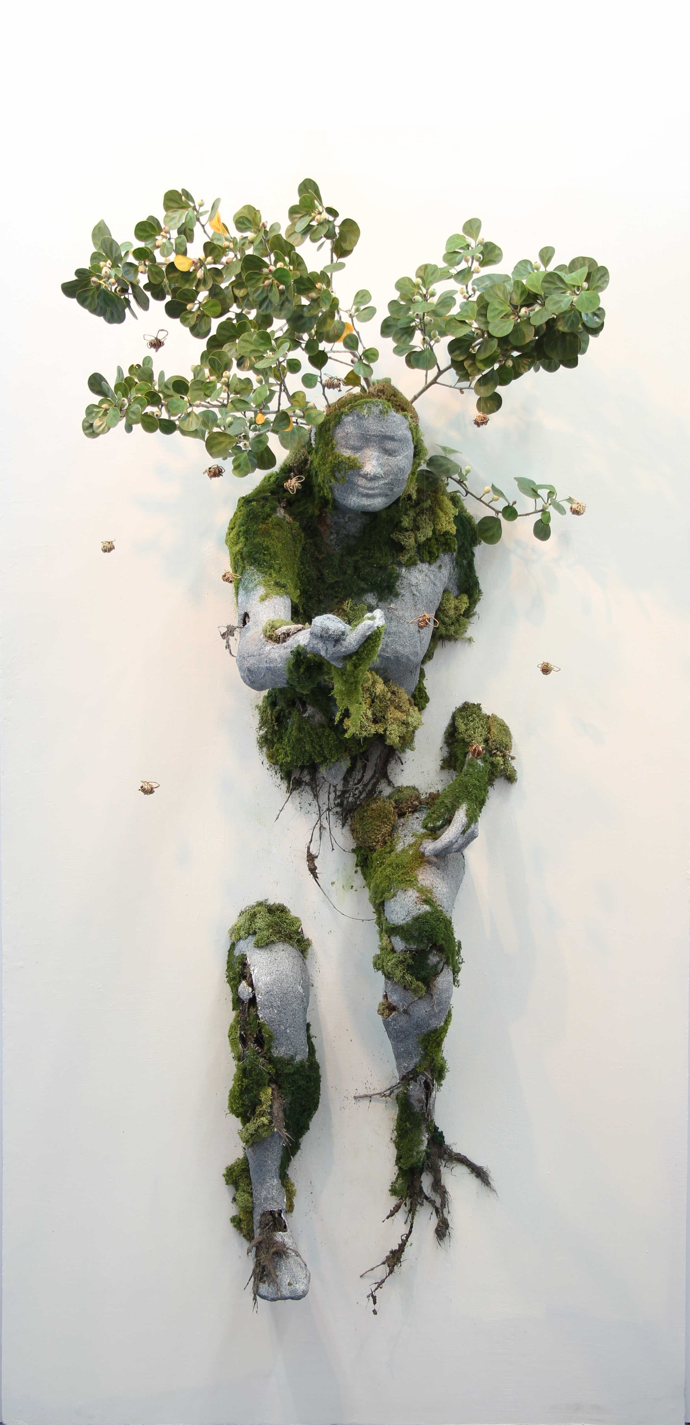 Photograph of a gray stone figure covered in green plant leaves and moss, hanging on a white wall