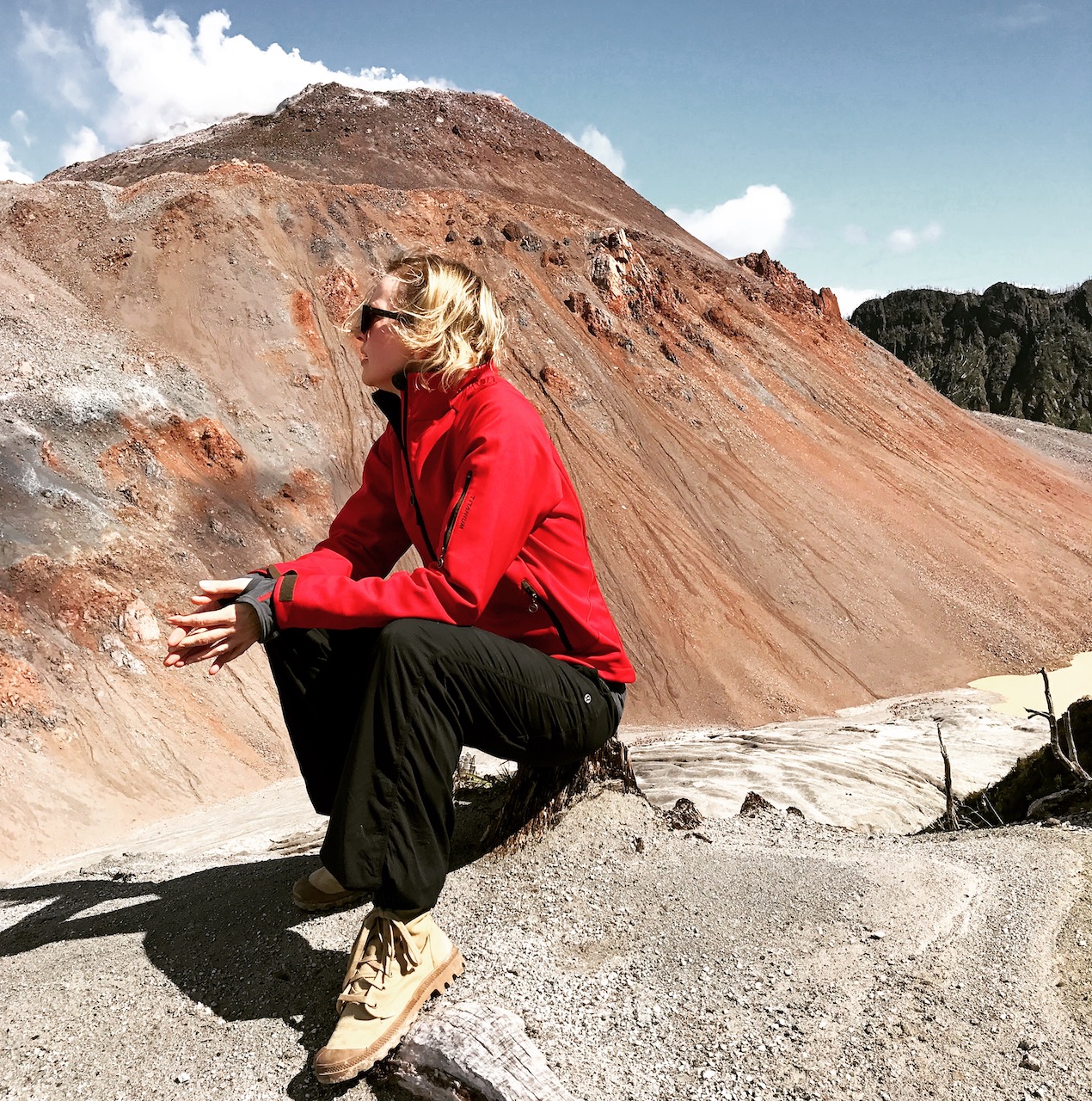 Photograph of a person with light-colored skin and shoulder length blonde hair wearing a red jacket, black pants, and tan hiking boots, sitting on a stump with a rock formation in the distance