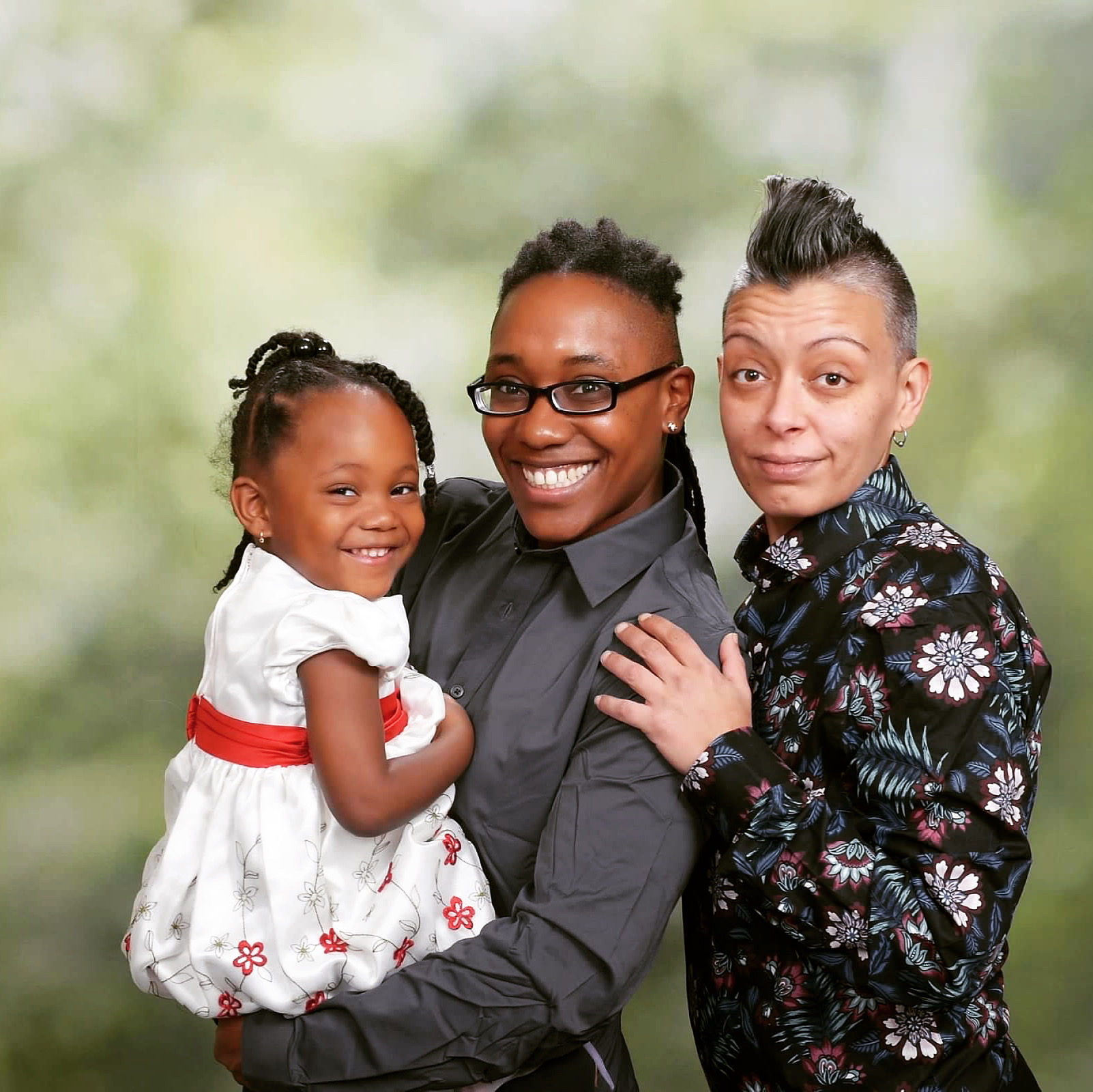 Photograph of three people against a greenish-white background, including a child with medium-colored skin wearing a white dress with a red belt, held by a person with medium-colored skin wearing a long-sleeved gray shirt, standing next to a person with light-colored skin wearing a long-sleeved black shirt with flowers