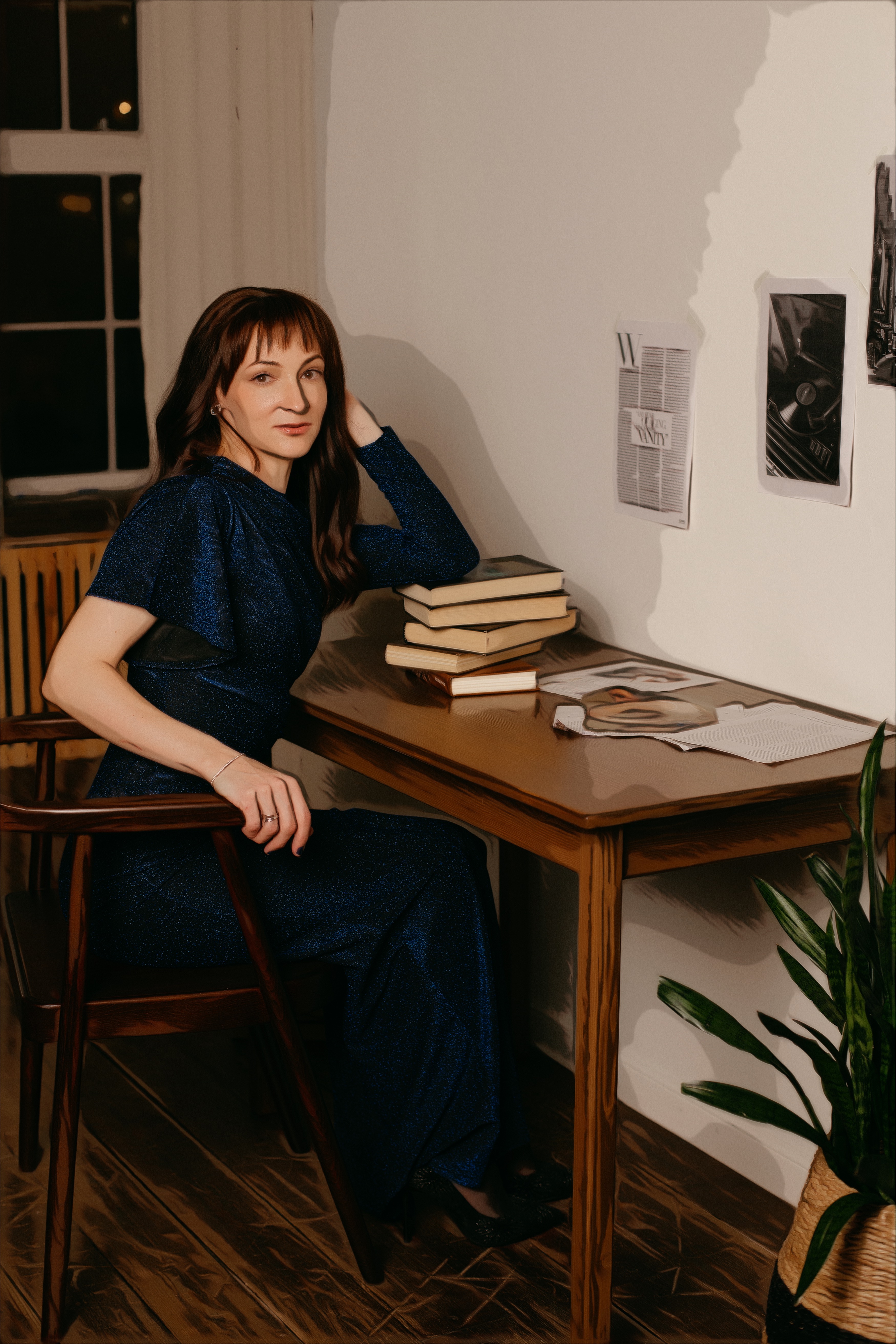 Anastasia, a tall woman with long dark hair and a fringe is sitting at a wooden desk, a stack of books is supporting one of her hands that props up her head.  Anastasia is looking at a camera smiling. She wears a gorgeous long blue sparkling dress with one long and one short sleeve showing her hand.