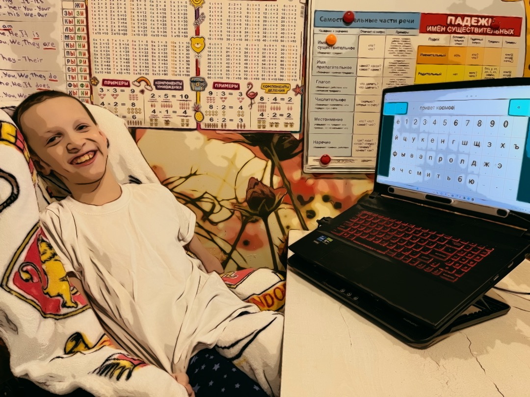 Ivan, a widely-smiling pale-skinned person with hazelnut eyes, is sitting in his chair covered by a light plush blanket. He sits in front of his desk and laptop which is open and shows a Russian alphabet and a typed phrase “hello cosmos.” A pale grey stripe of the eyetracker is attached between the upper and lower parts of the laptop. Behind Ivan, on the wall, there are a lot of posters with school rules from Math, Russian, and English. The wallpaper peeking through the posters shows beautiful red flowers scratched a little bit at the bottom by cat’s claws.