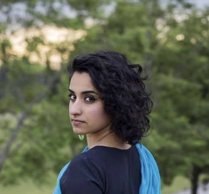 Photograph of a person with medium-colored skin and shoulder length brown hair, wearing a black shirt with a blue scarf over one shoulder, against a background of trees and a sunset