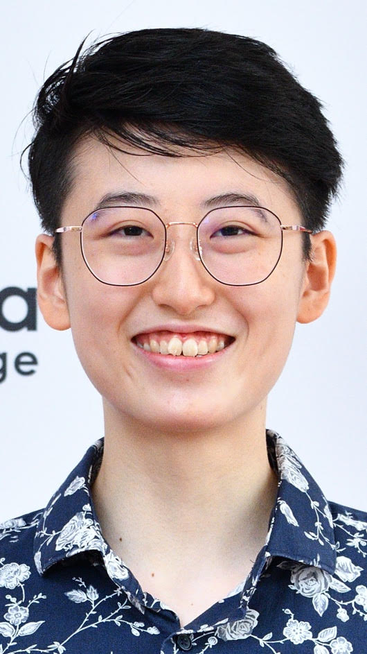 Photograph of a person with light-colored skin and dark hair wearing wire eyeglasses and a dark blue shirt with white flowers, against a white background