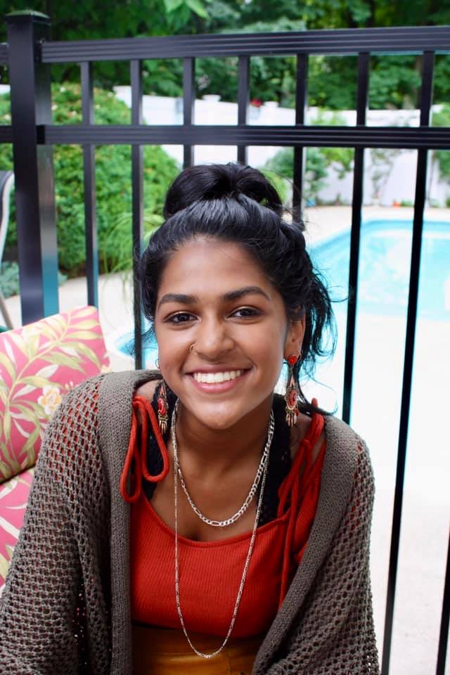 Photograph of person with black hair pulled up in a bun, medium brown skin, wearing a crocheted brown sweater and a red shirt