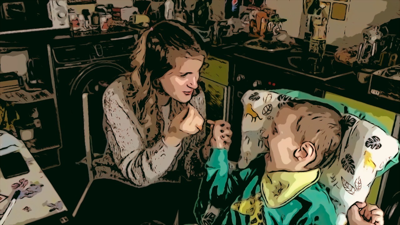 Ivan’s teacher and Ivan sit side by side, turned towards each other. Both smile, looking at each other. His teacher has long blonde hair tied into two little ponytails on top of her head, the rest of the hair is flowing loose down in front of her grey sweater. Ivan is wearing a green shirt with giraffe and a light green bib on his neck. The teacher holds Ivan’s hand with one hand and emotionally gesticulates with the other.