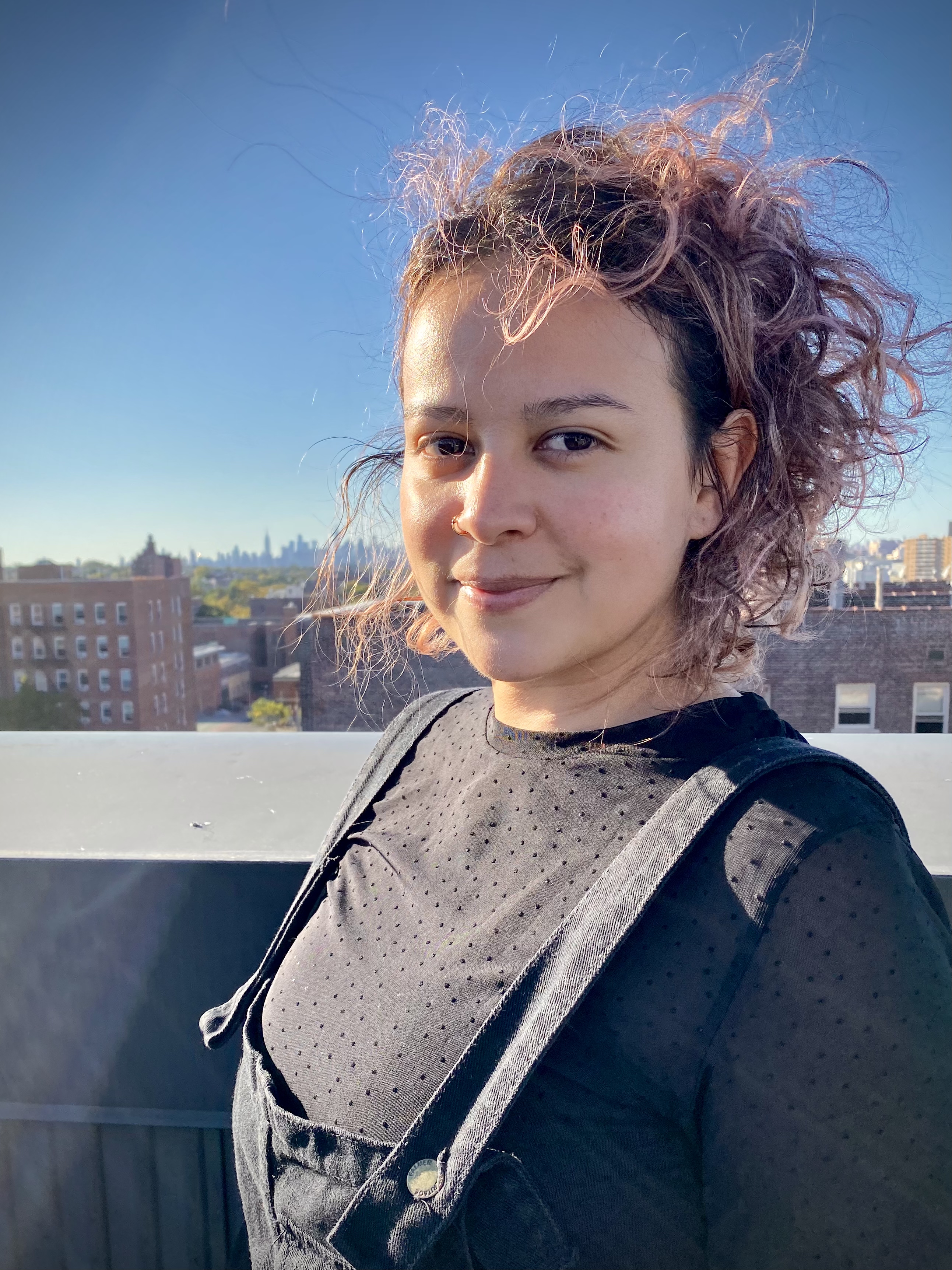 Photograph of a person with light-colored skin and chin-length pink hair, wearing a black shirt and black overalls, standing on a rooftop with a city skyline in the background