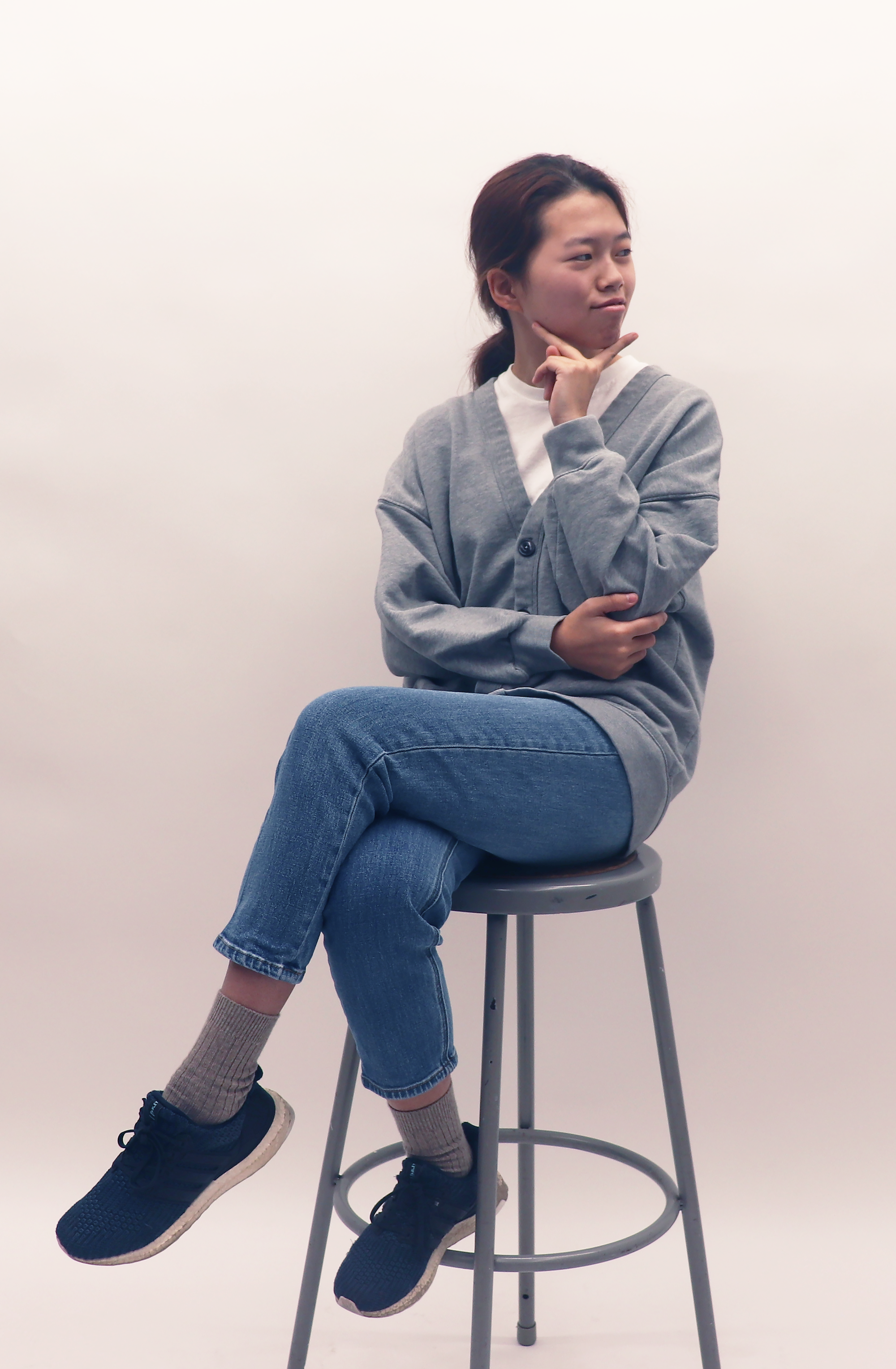 Photograph of a person with light-colored skin and dark hair pulled back, wearing gray sweater, white shirt, blue jeans, gray socks, and black sneakers, sitting on a gray metal stool against a white background