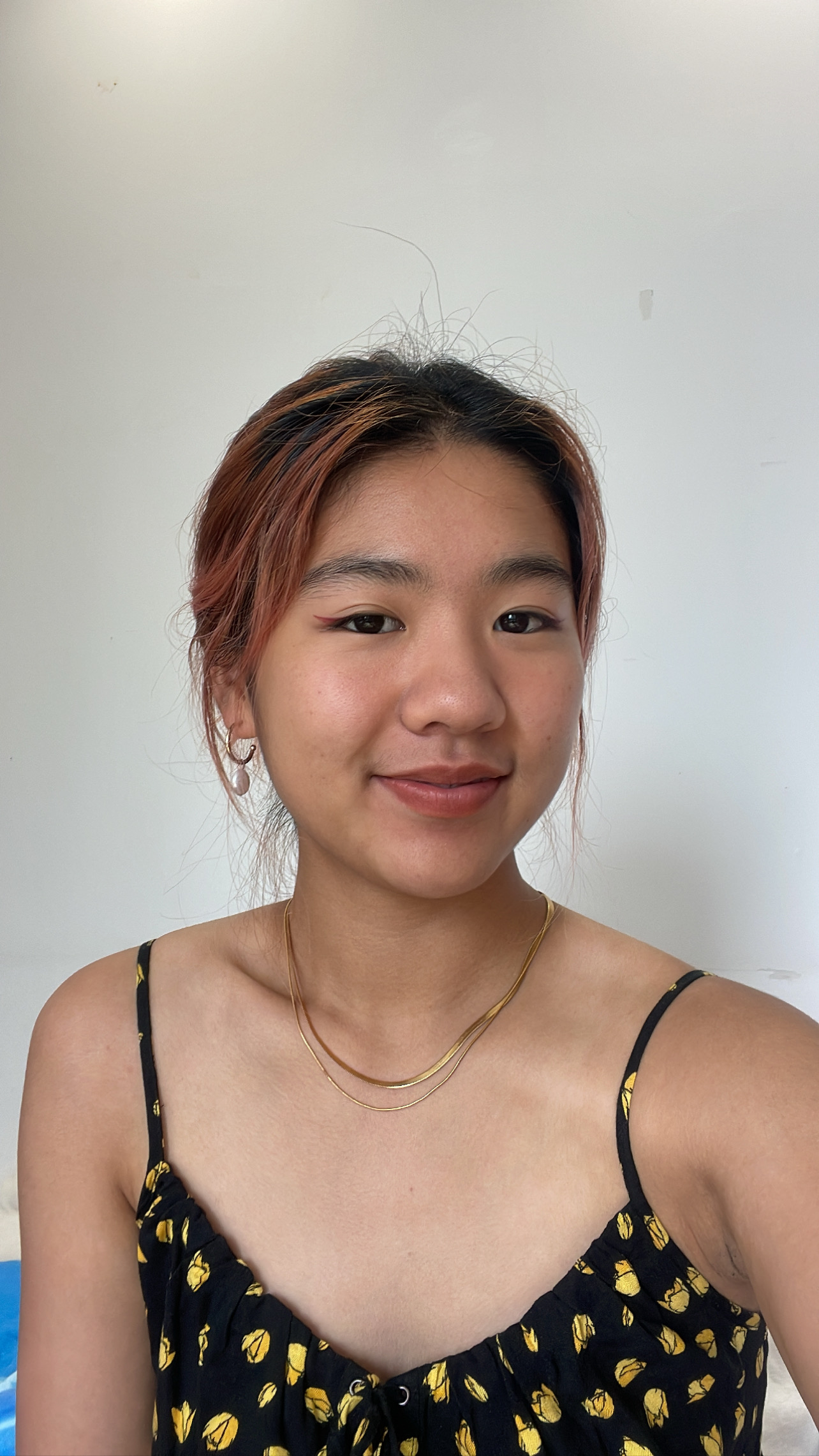 Photograph of person with reddish brown and black hair pulled back, light beige skin, wearing a black and yellow printed shirt