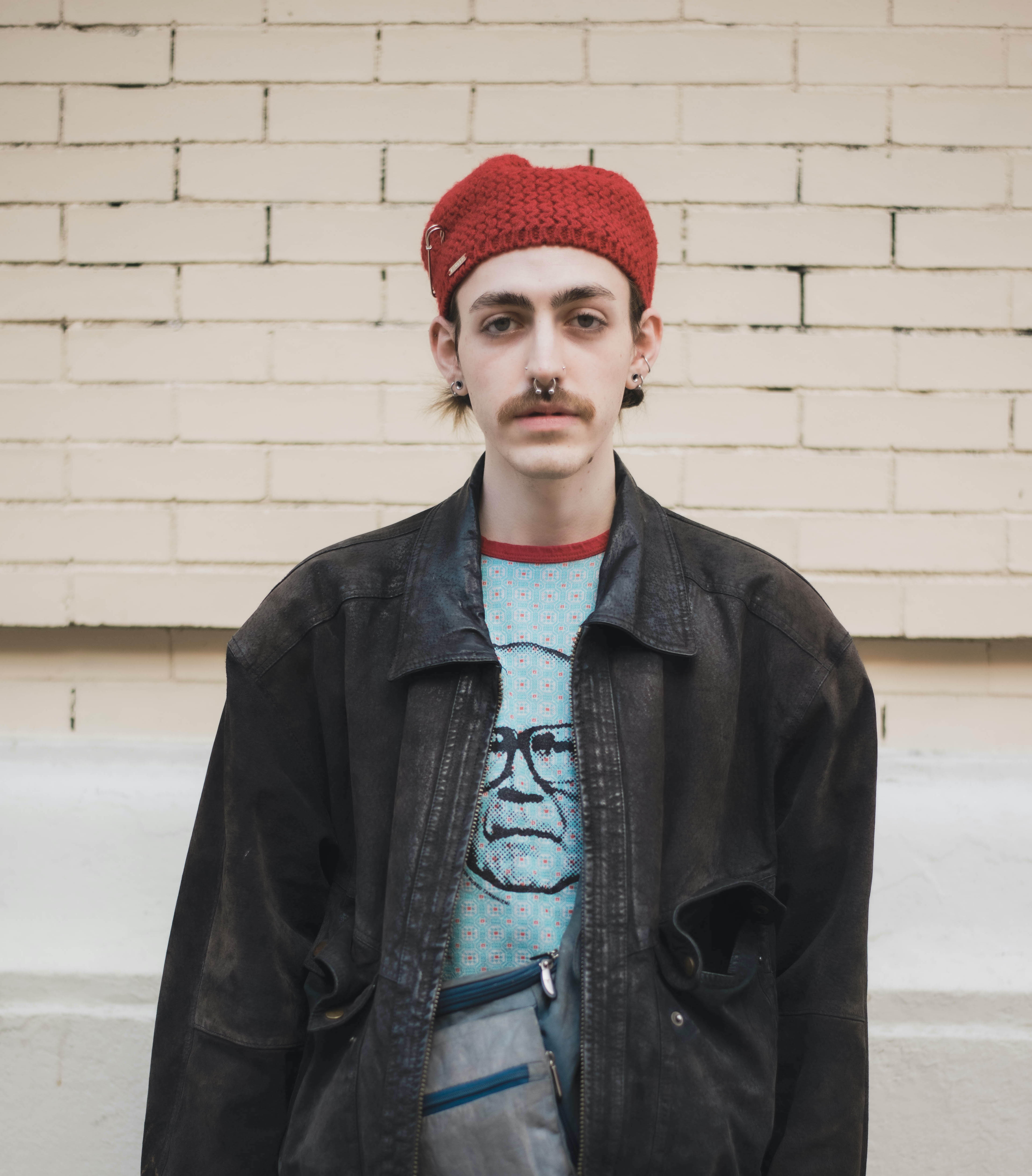 Photograph of person with crocheted red beret, light beige skin, wearing dark gray jacket and light blue patterned t-shirt with red collar