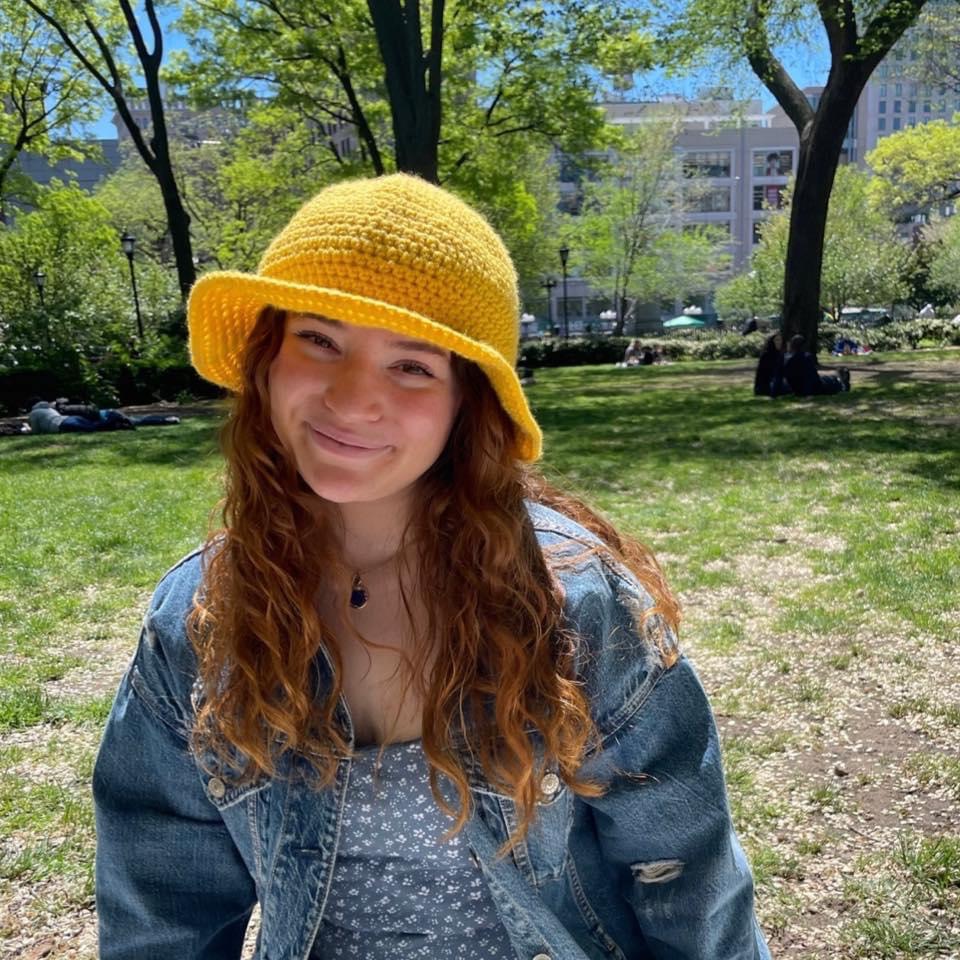 Photograph of person with light reddish brown hair and a crocheted yellow hat, light beige skin, wearing a blue jacket and blue and white printed shirt