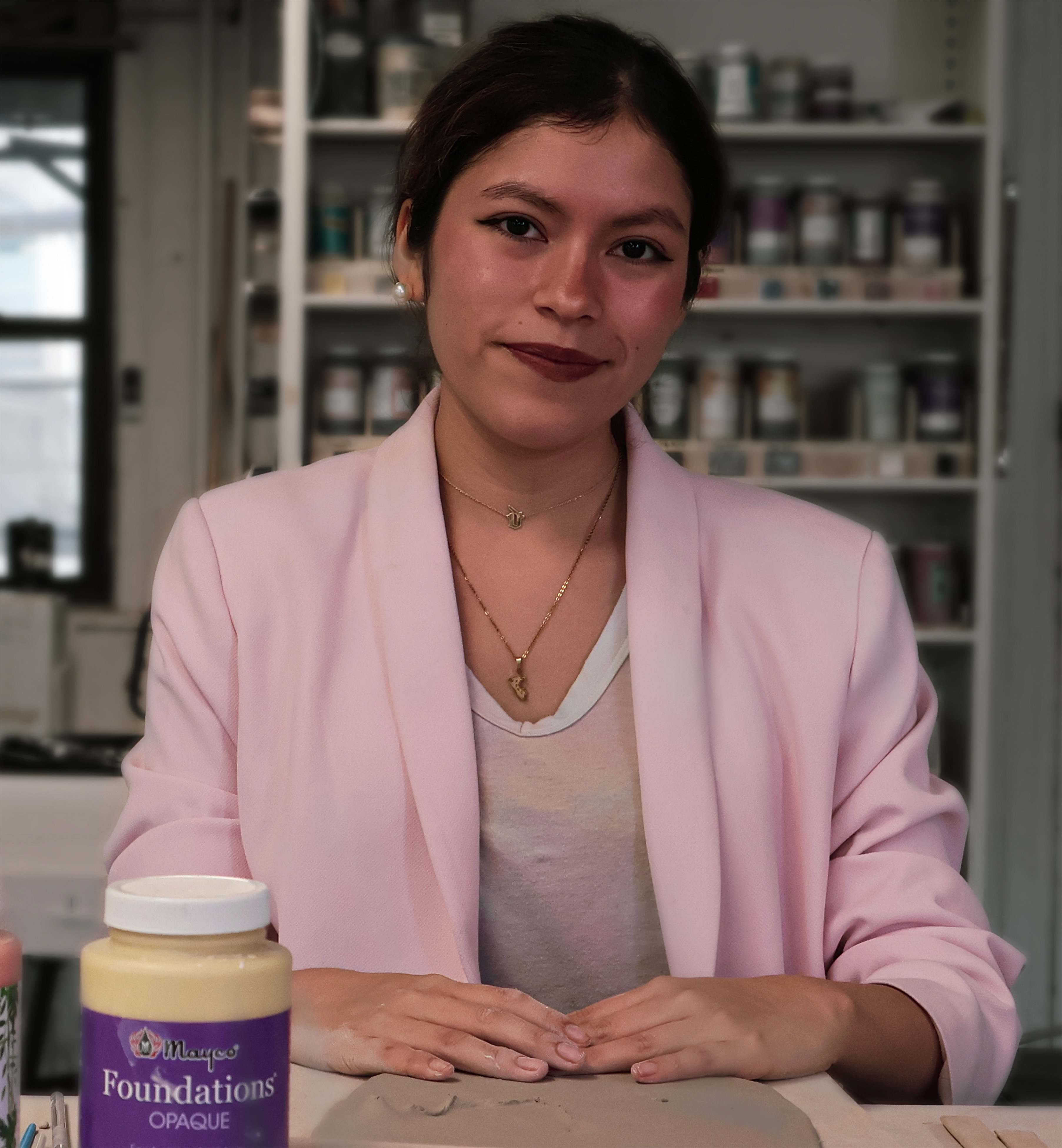 Photograph of a person with light-colored skin and dark hair pulled back, wearing a pink blazer and tan shirt with two gold necklaces, sitting in a room filled with art supplies