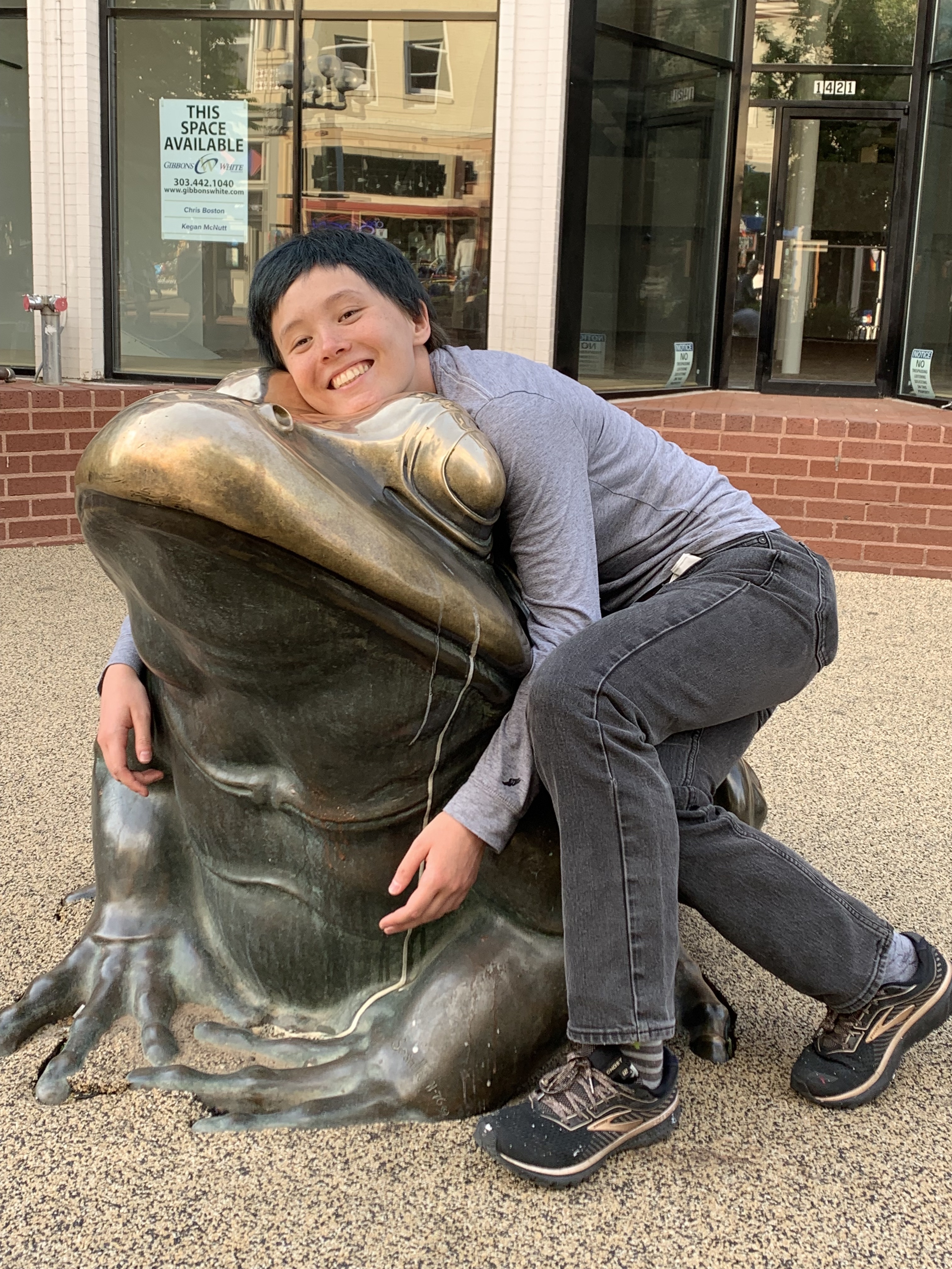 Photograph of a person with light-colored skin, short dark hair, wearing a gray shirt and black jeans, leaning against a large bronze frog sculpture