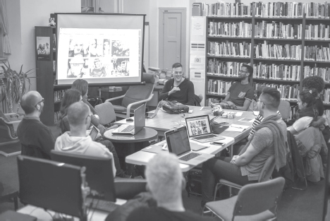 People sitting in a library talking and looking at a projector featuring images of drag queens in Canada.