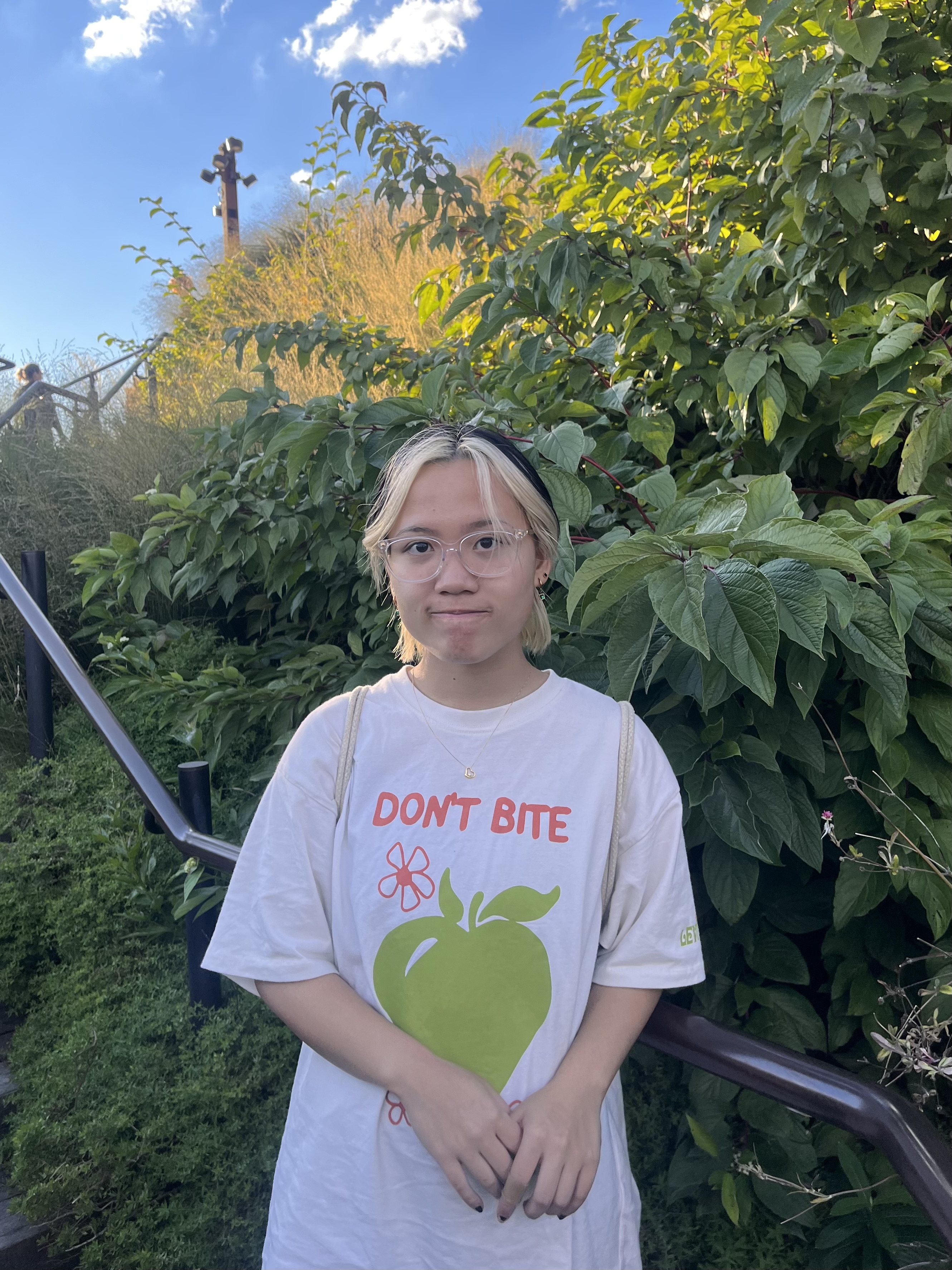 Photograph of a person with light-colored skin and light-dark hair wearing white t-shirt with writing and a drawing of a green apple, standing against a background of green leaves