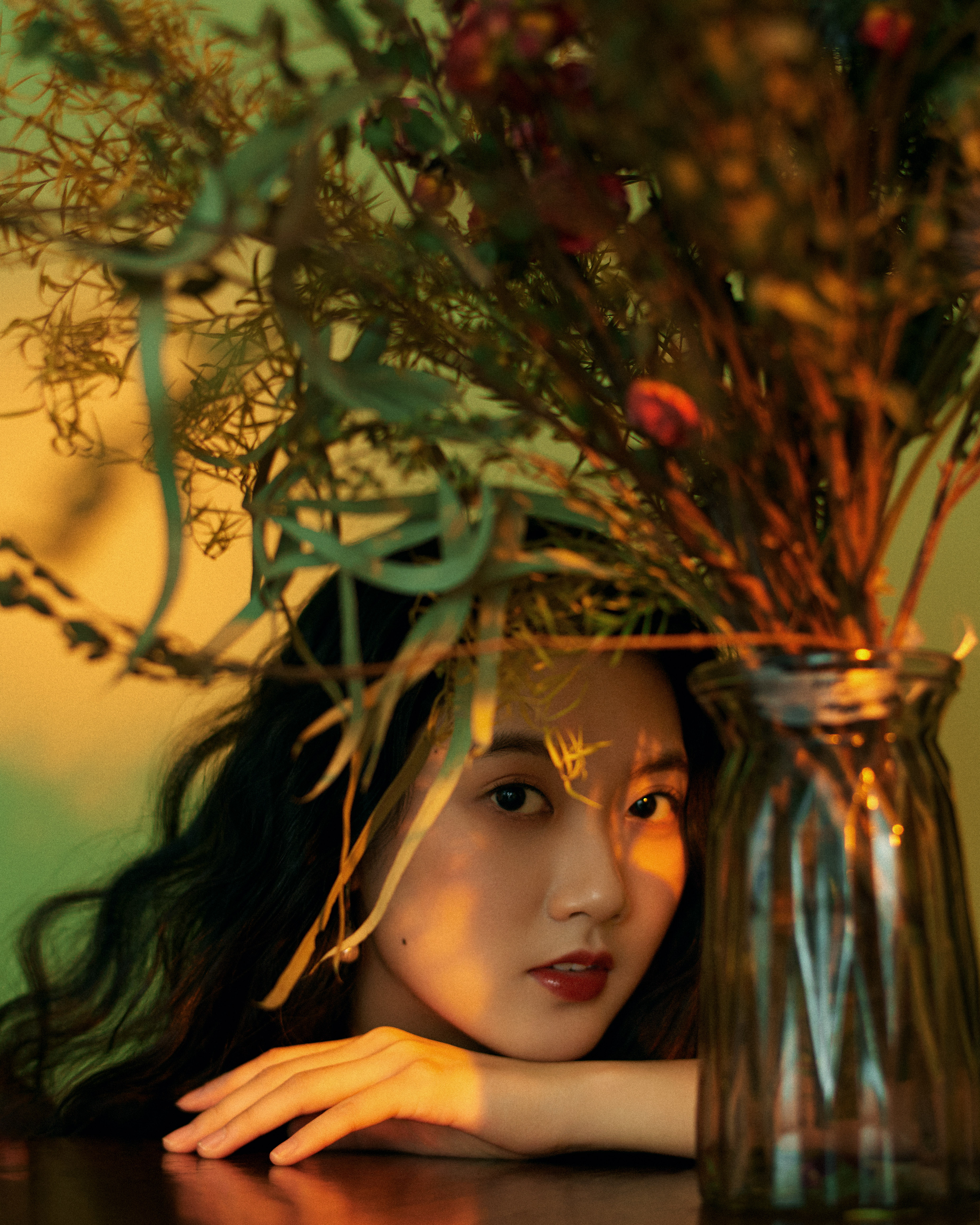 Photograph of a person with light-colored skin and long dark hair, posed behind a vase of dried flowers and leaf stalks