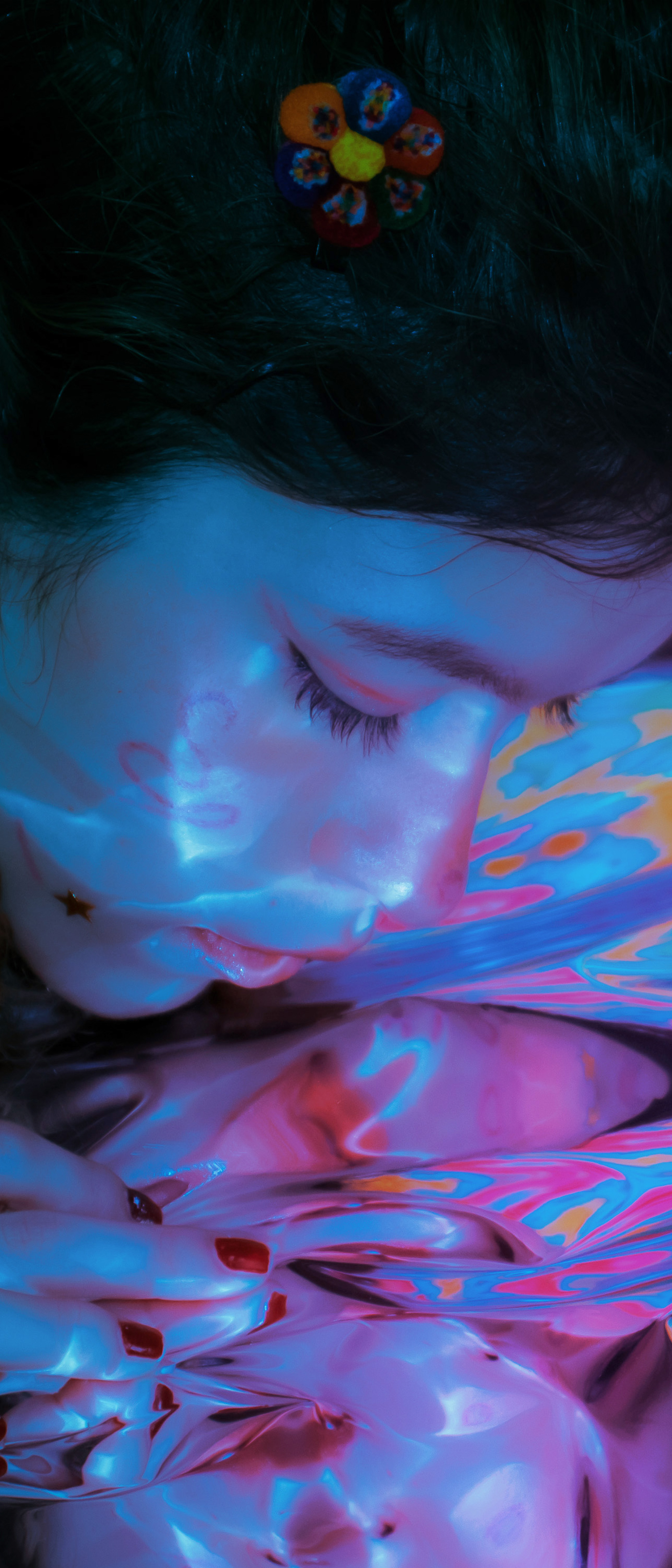 Close-up photograph of a person wearing a flower hair decoration, looking into a mirrored reflection, highlighted in black, blues, pinks, orange, reds