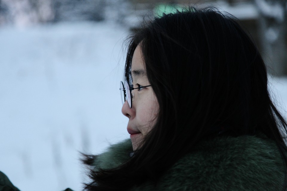 Photograph of a person with light-colored skin and dark hair wearing wire eyeglasses, staring to the left against a blurred white background