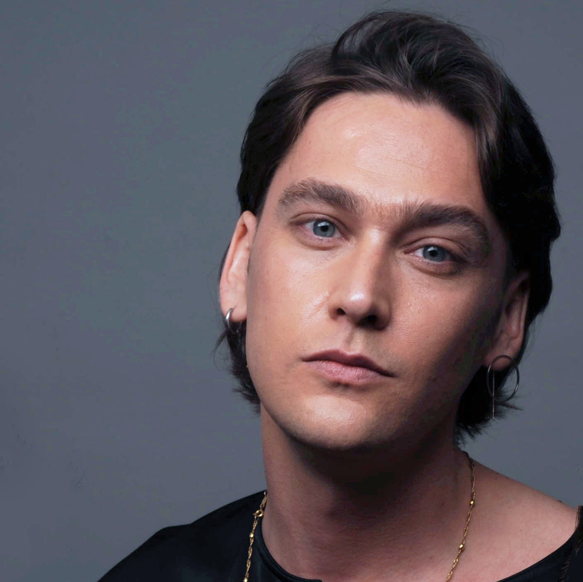 Photograph of a person with light-colored skin and short brown hair, wearing a black shirt and gold jewelry, against a gray background