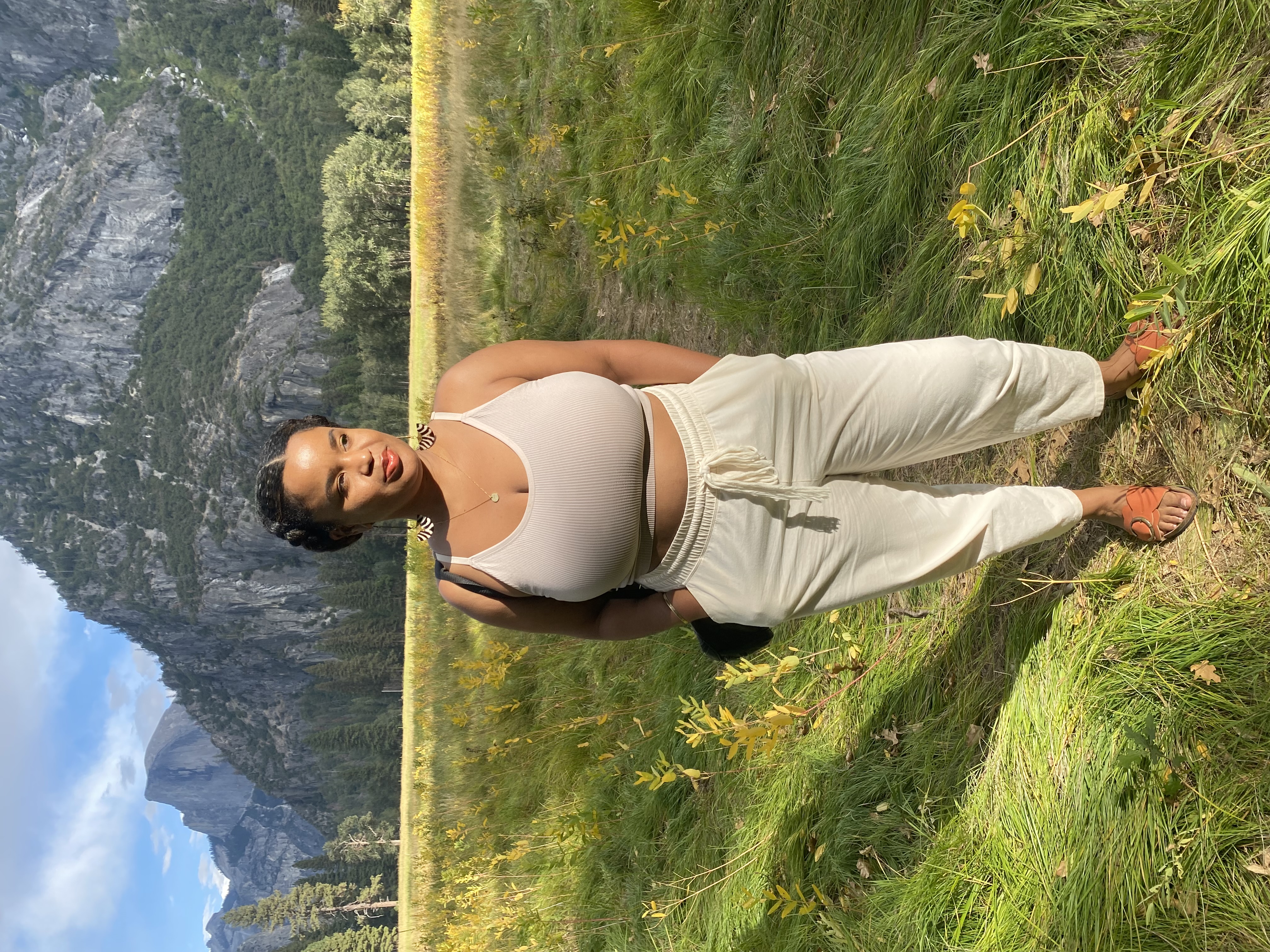 Photograph of a person with medium-colored skin and dark hair in French braids, wearing a beige tank top and and cream-colored pants, standing on green grass in front of a gray mountain