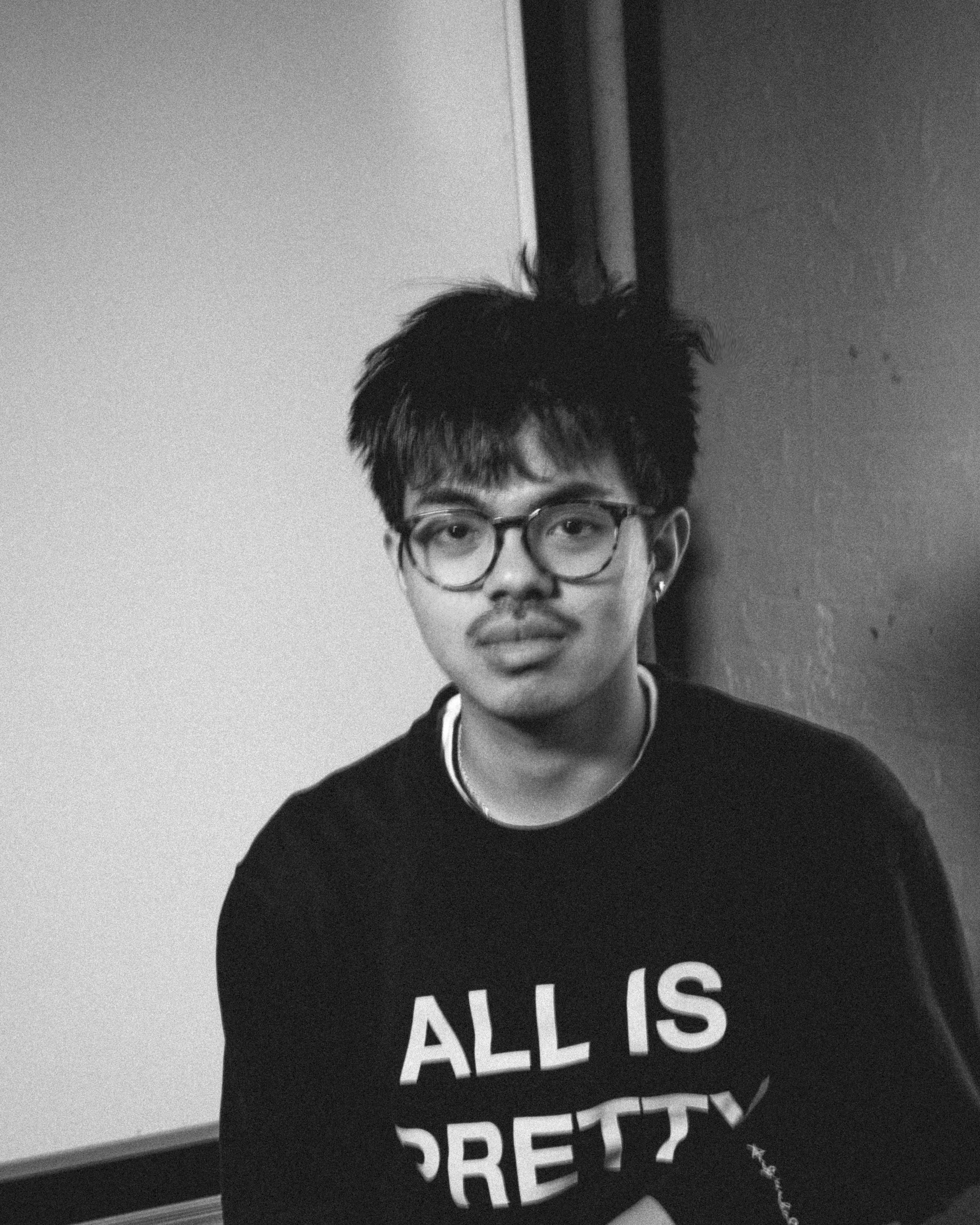 Black and white photograph of person with black hair, light beige skin, and glasses, wearing a black sweatshirt with white letters