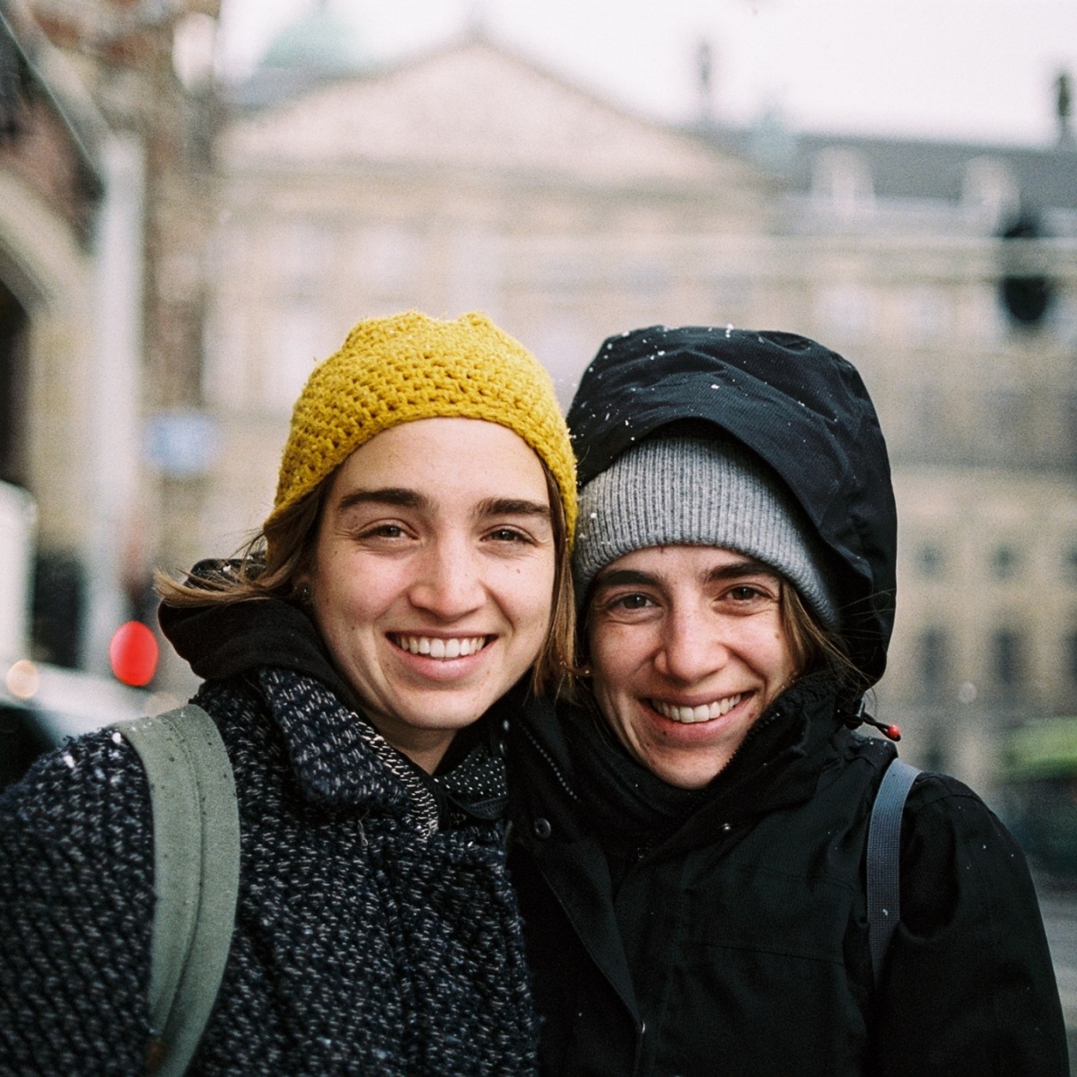 Photograph of two people with light-colored skin and brown hair, one wearing a yellow crocheted skullcap and black-gray patterned coat and the other wearing a knit gray skullcap and a black coat and hood